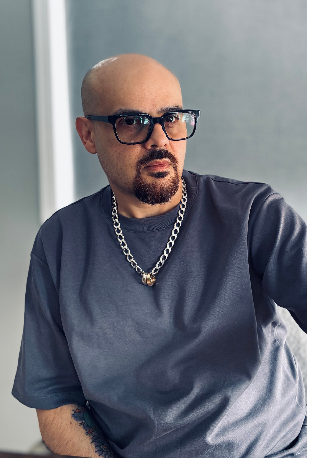 Miguel with glasses and silver chain necklace wearing a gray t-shirt, looking downward.